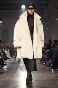 Model on a runway wearing an oversized white puffer coat over a black turtleneck and leather skirt, with a black futuristic visor.