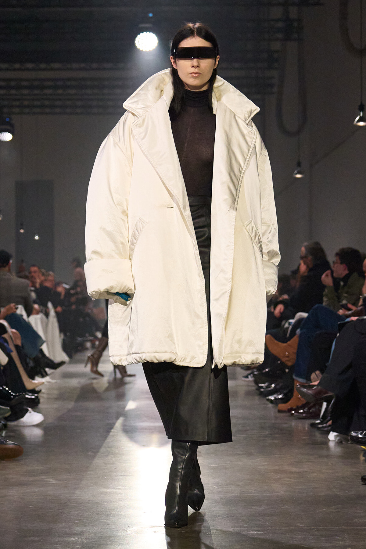 Model on a runway wearing an oversized white puffer coat over a black turtleneck and leather skirt, with a black futuristic visor.
