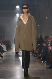 Model walks a runway in an oversized taupe ribbed zip-front cardigan over a white shirt, black pants, boots, and a futuristic black visor.