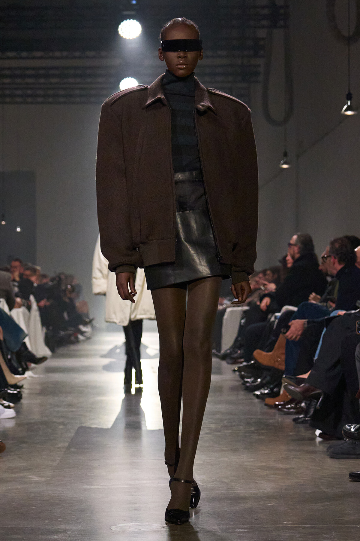 Model on a runway wearing an oversized brown jacket over a black turtleneck, leather mini skirt, sheer tights, black ankle-strap heels, and a rectangular black eyewear/face shield.