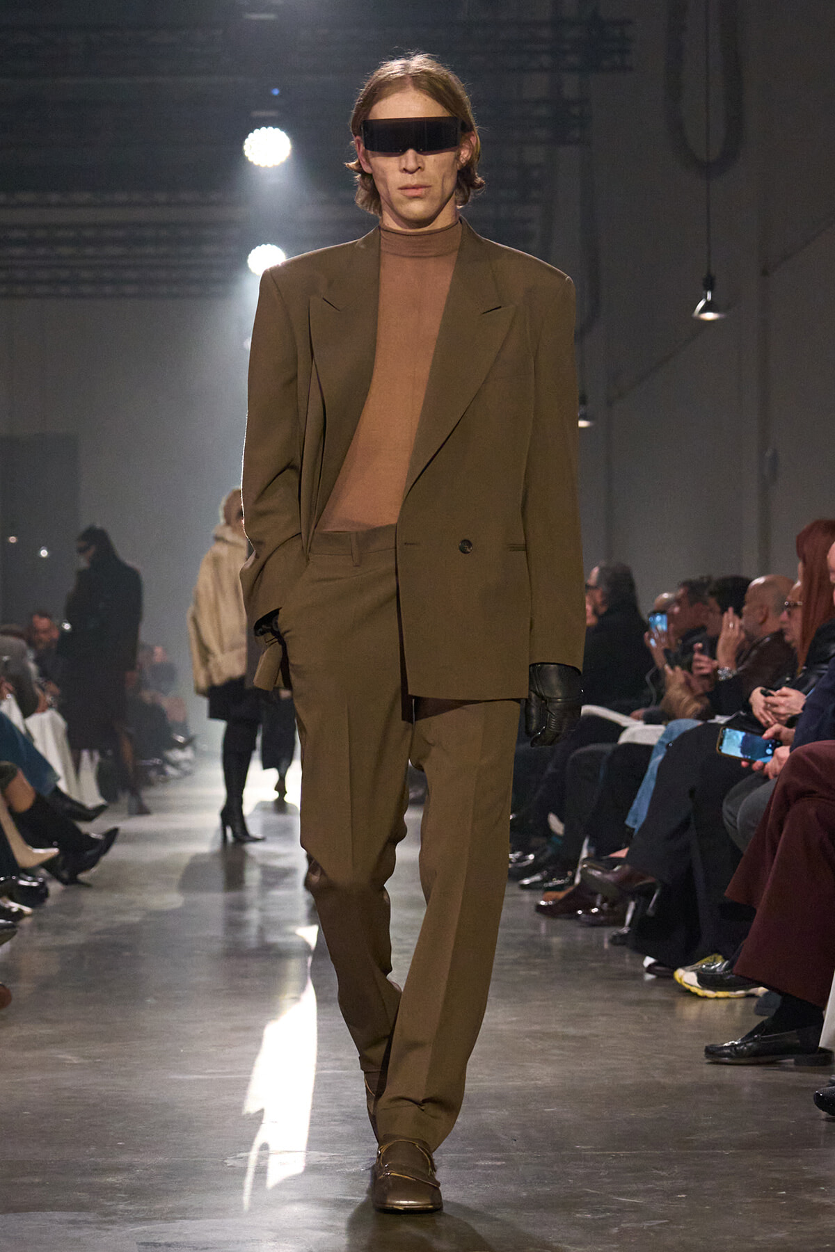 Male model wearing a brown suit with a brown turtleneck, black gloves, and futuristic black sunglasses on a fashion runway.