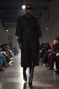 Model walks the runway in an all-black look: oversized belted trench coat, black leather gloves, knee-high boots, and a wide black visor-style sunglasses; audience in the background.
