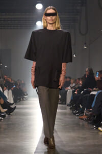 Model walks a fashion show runway in an oversized black top, gray trousers, and pink gloves, wearing a futuristic dark visor sunglasses.