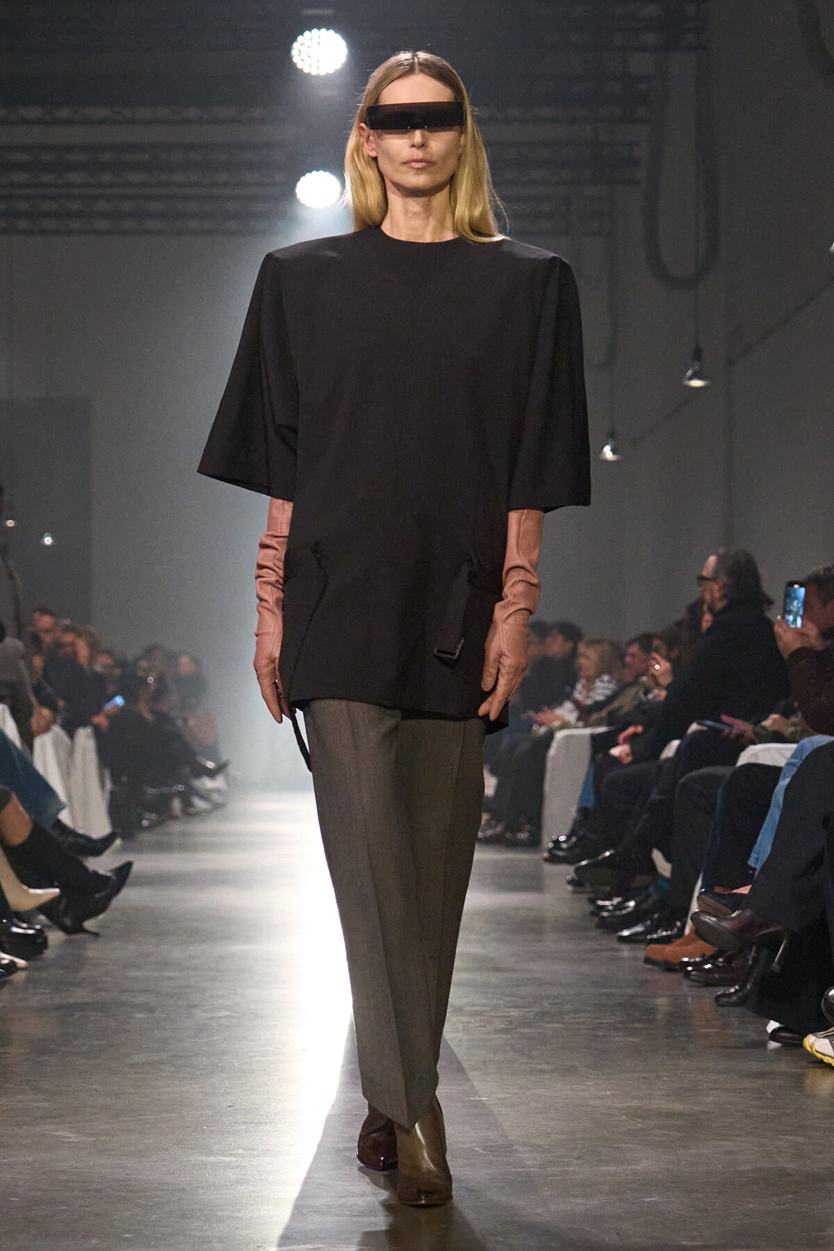 Model walks a fashion show runway in an oversized black top, gray trousers, and pink gloves, wearing a futuristic dark visor sunglasses.