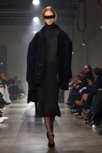 Model walks a fashion show wearing an oversized dark coat over a black turtleneck dress with a futuristic rectangular visor-style sunglasses and black heels.