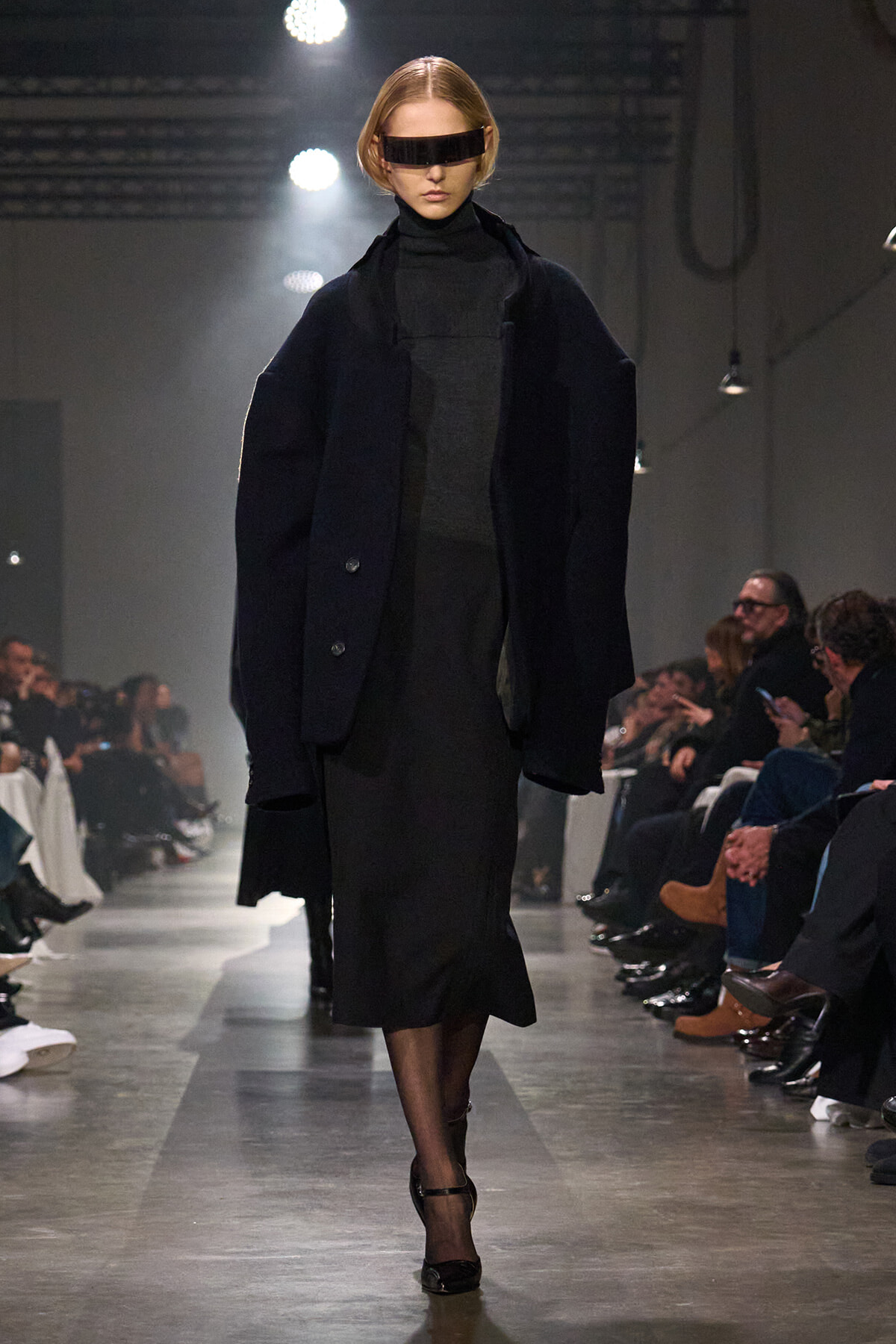 Model walks a fashion show wearing an oversized dark coat over a black turtleneck dress with a futuristic rectangular visor-style sunglasses and black heels.