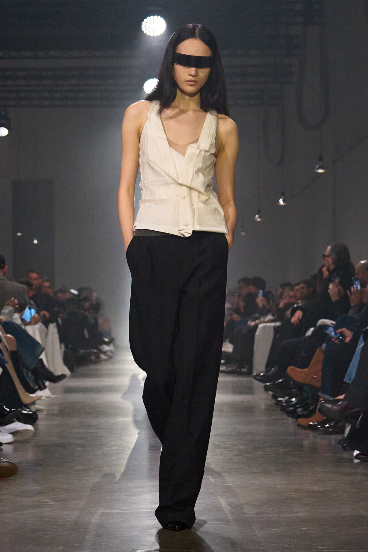 Model walking a fashion runway in a cream sleeveless top and black wide-leg pants, with a black blindfold across her eyes.