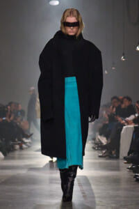 Runway model in an oversized black coat over a turquoise dress and black knee-high boots, wearing a rectangular black visor accessory.