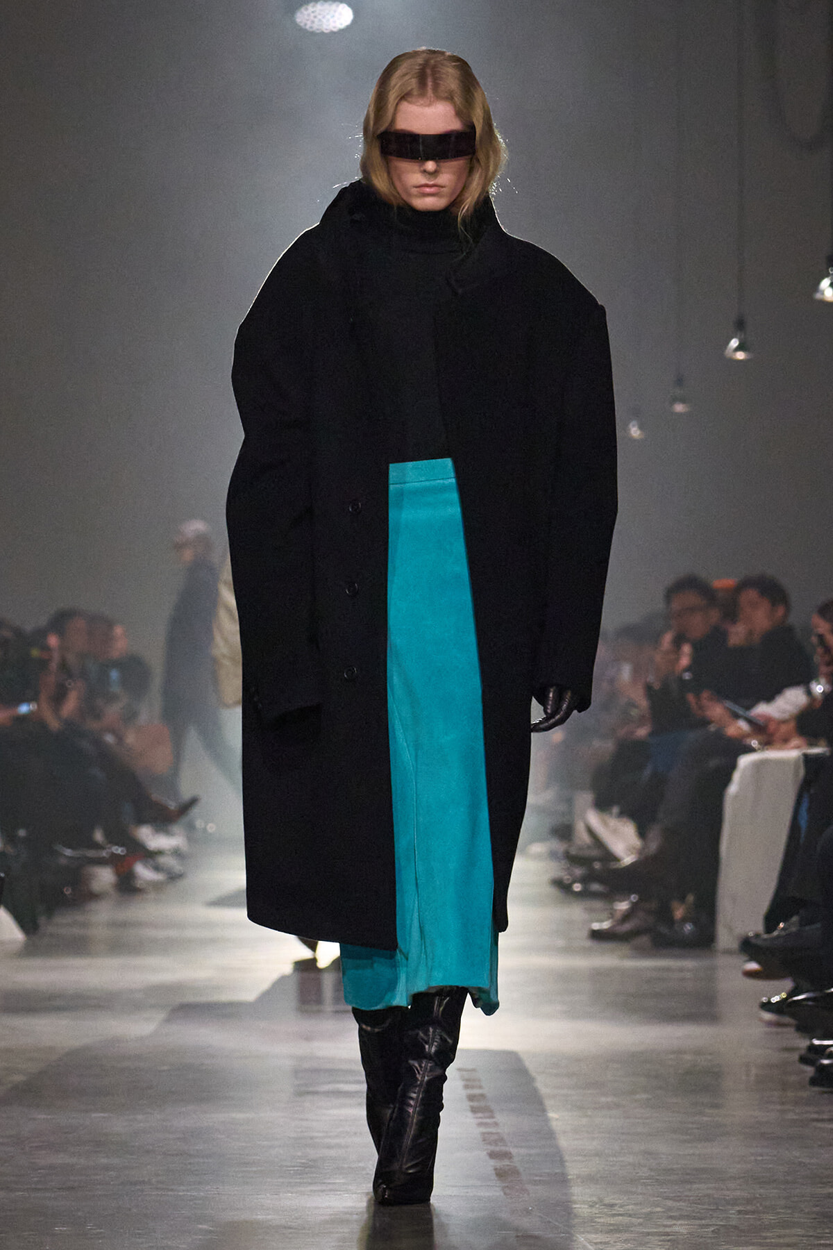 Runway model in an oversized black coat over a turquoise dress and black knee-high boots, wearing a rectangular black visor accessory.