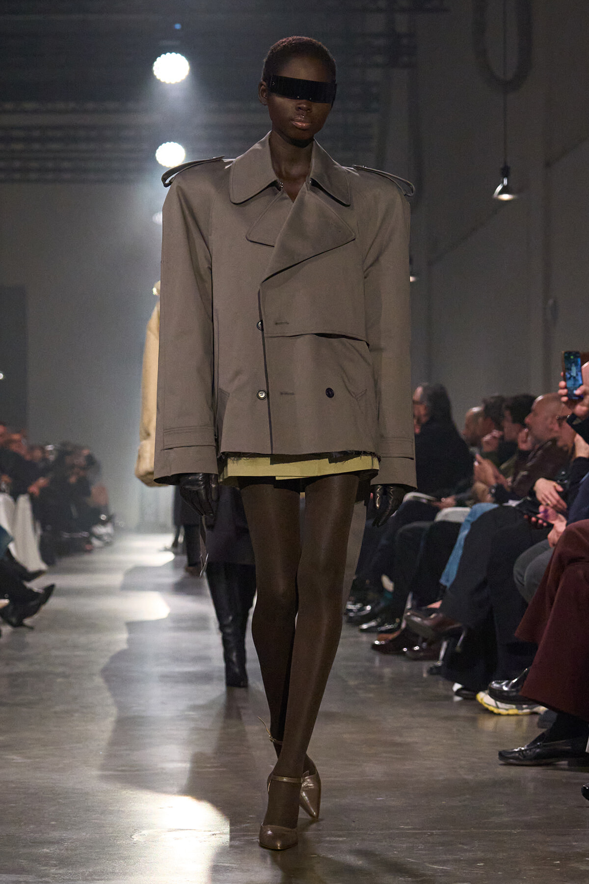 Model on a runway wearing an oversized beige trench coat, black visor sunglasses, gloves, and tan heels among seated audiences.