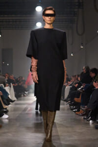Model in a black knee-length dress with wide short cape sleeves, brown elbow-length gloves, tan tall boots, and futuristic visor sunglasses on a runway.