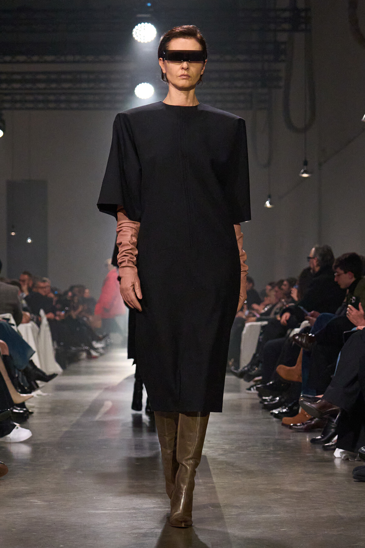 Model in a black knee-length dress with wide short cape sleeves, brown elbow-length gloves, tan tall boots, and futuristic visor sunglasses on a runway.