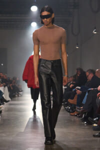 Male model on a runway wearing a skin-toned fitted top, black leather pants, and a large black visor sunglasses.
