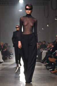 Model walks the runway in a sheer black turtleneck top and wide-leg black trousers, wearing a futuristic visor over her eyes.