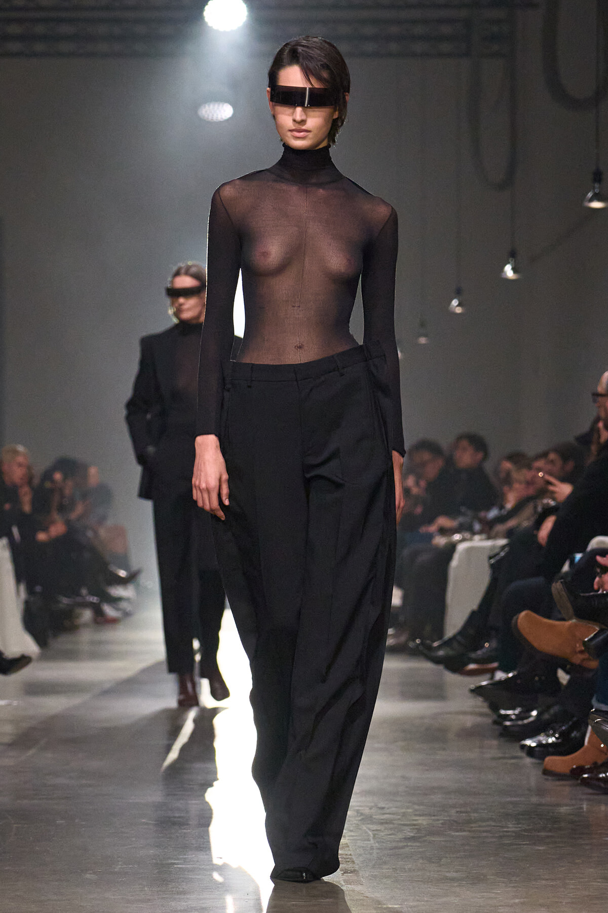 Model walks the runway in a sheer black turtleneck top and wide-leg black trousers, wearing a futuristic visor over her eyes.
