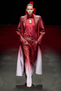 Model on a runway wearing a red gradient pantsuit with a long burgundy coat and a gold pendant necklace, hands in pockets.
