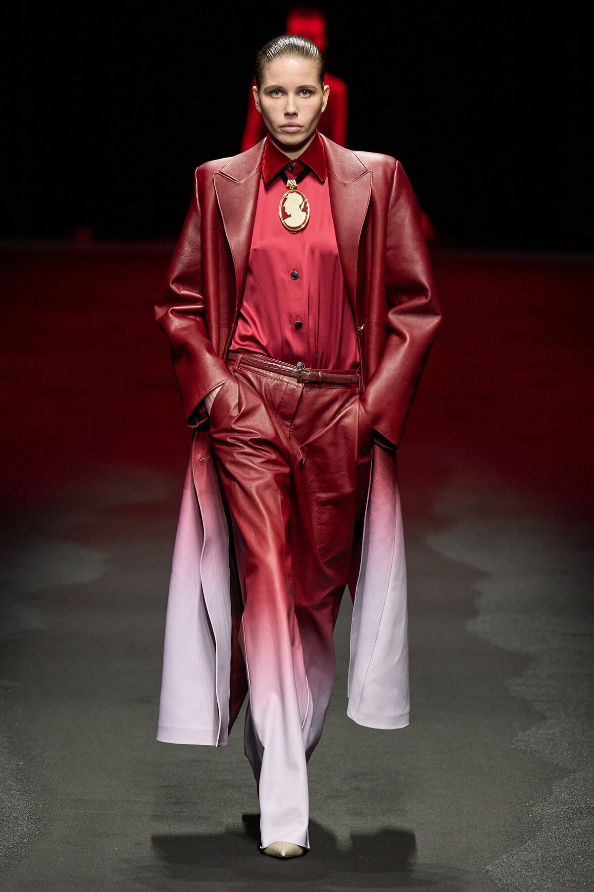 Model on a runway wearing a red gradient pantsuit with a long burgundy coat and a gold pendant necklace, hands in pockets.