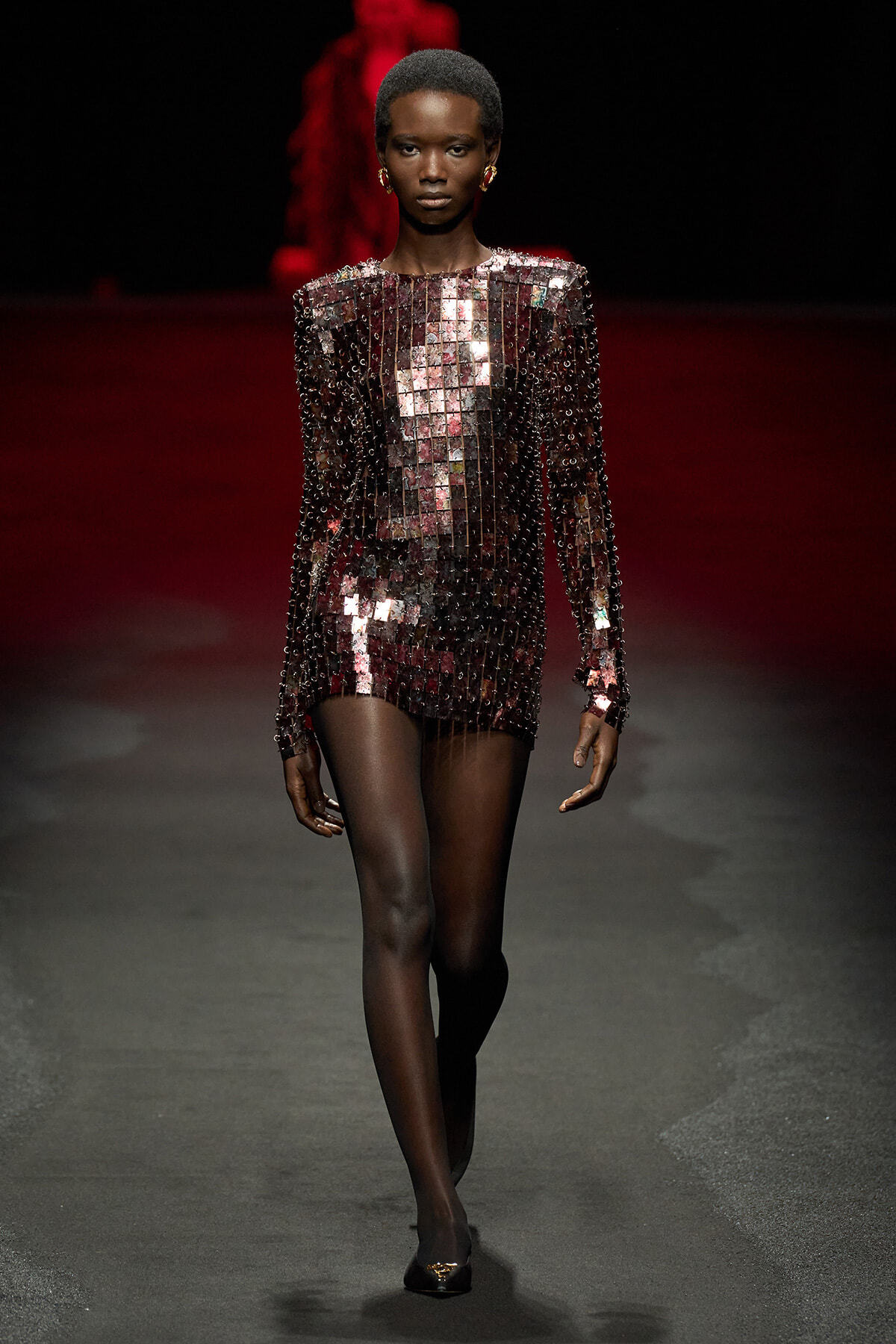 Model walking a fashion show in a burgundy sequined mini dress with long sleeves and square mirrored panels under red-tinged lights on a dark runway. Runway model in a sparkly burgundy dress covered in square mirrored sequins, long sleeves, gold earrings, on a dim catwalk. A model showcases a burgundy rhinestone dress composed of square mirrored tiles during a runway show. Model on a runway wearing a burgundy sequin mini dress with reflective square panels and sheer tights.