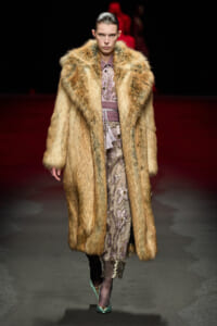 Model walks the runway in a long tan fur coat over a lilac patterned dress, paired with teal heels for a high-fashion look.