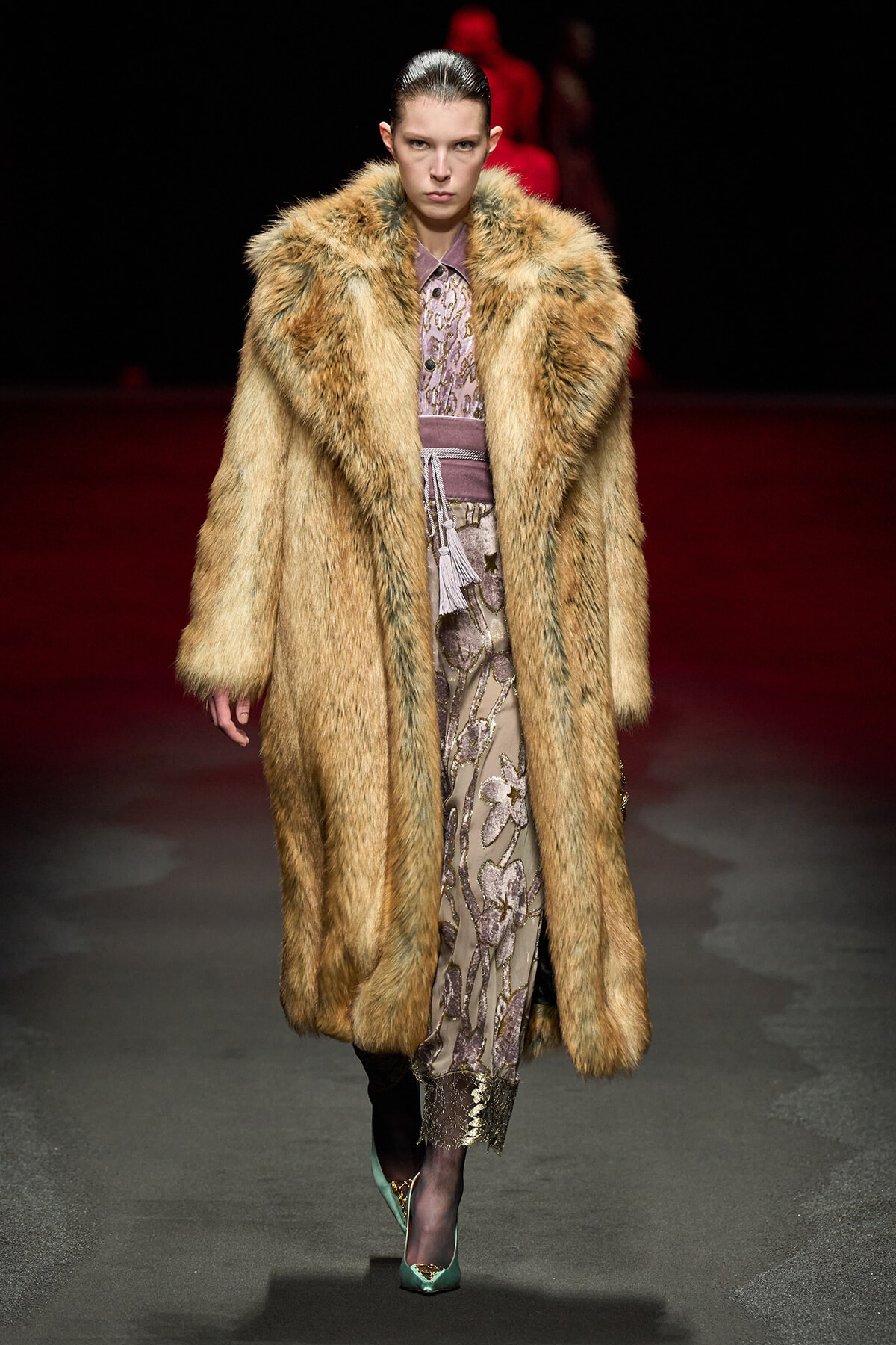 Model walks the runway in a long tan fur coat over a lilac patterned dress, paired with teal heels for a high-fashion look.