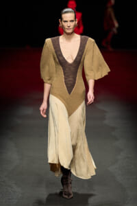 Model walking a fashion runway wearing a color-block, V-neck gown with brown and beige panels and dramatic puff sleeves, paired with dark boots.