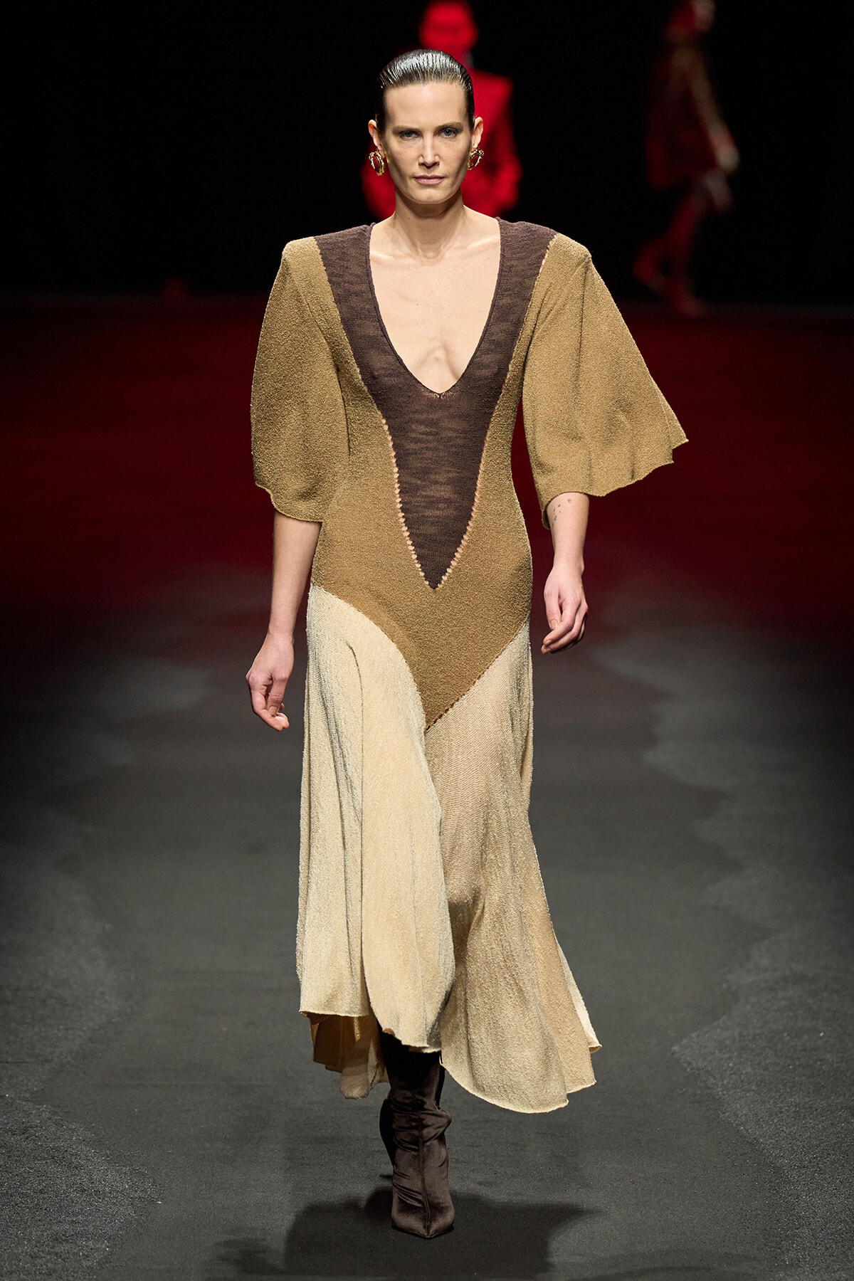 Model walking a fashion runway wearing a color-block, V-neck gown with brown and beige panels and dramatic puff sleeves, paired with dark boots.
