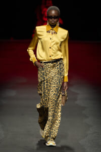 Model walks the runway in a yellow satin button-down shirt with leopard-print wide-leg pants, bold jewelry, and dark sunglasses.
