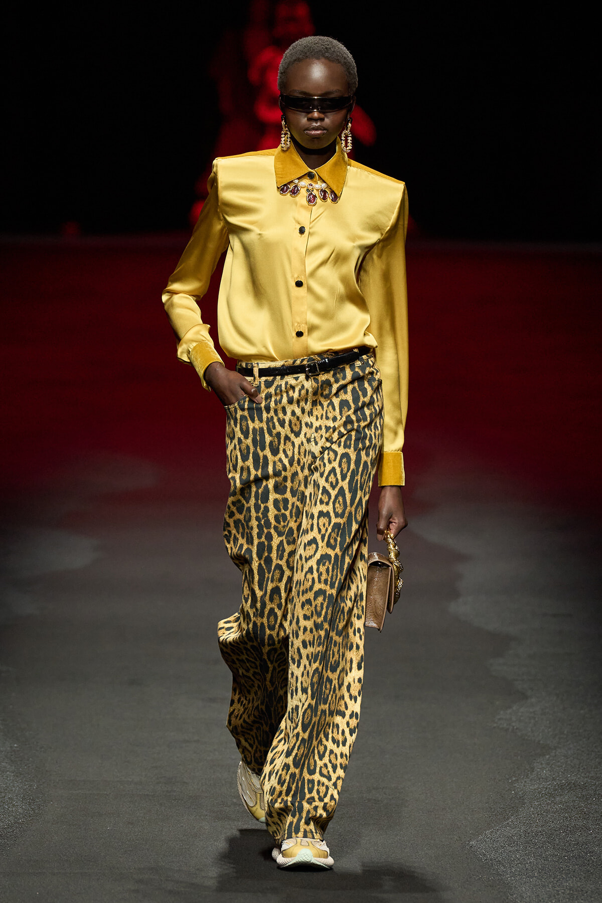Model walks the runway in a yellow satin button-down shirt with leopard-print wide-leg pants, bold jewelry, and dark sunglasses.