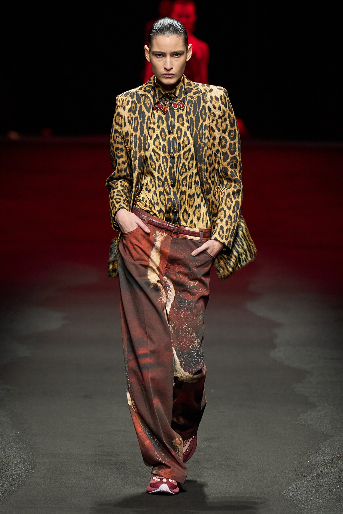 Model on a runway wearing a leopard-print blazer with a matching shirt and wide rust-colored printed pants.
