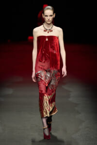 Model on a runway wearing a strapless red velvet dress with a draped, metallic gold-red skirt and a statement red gemstone necklace.