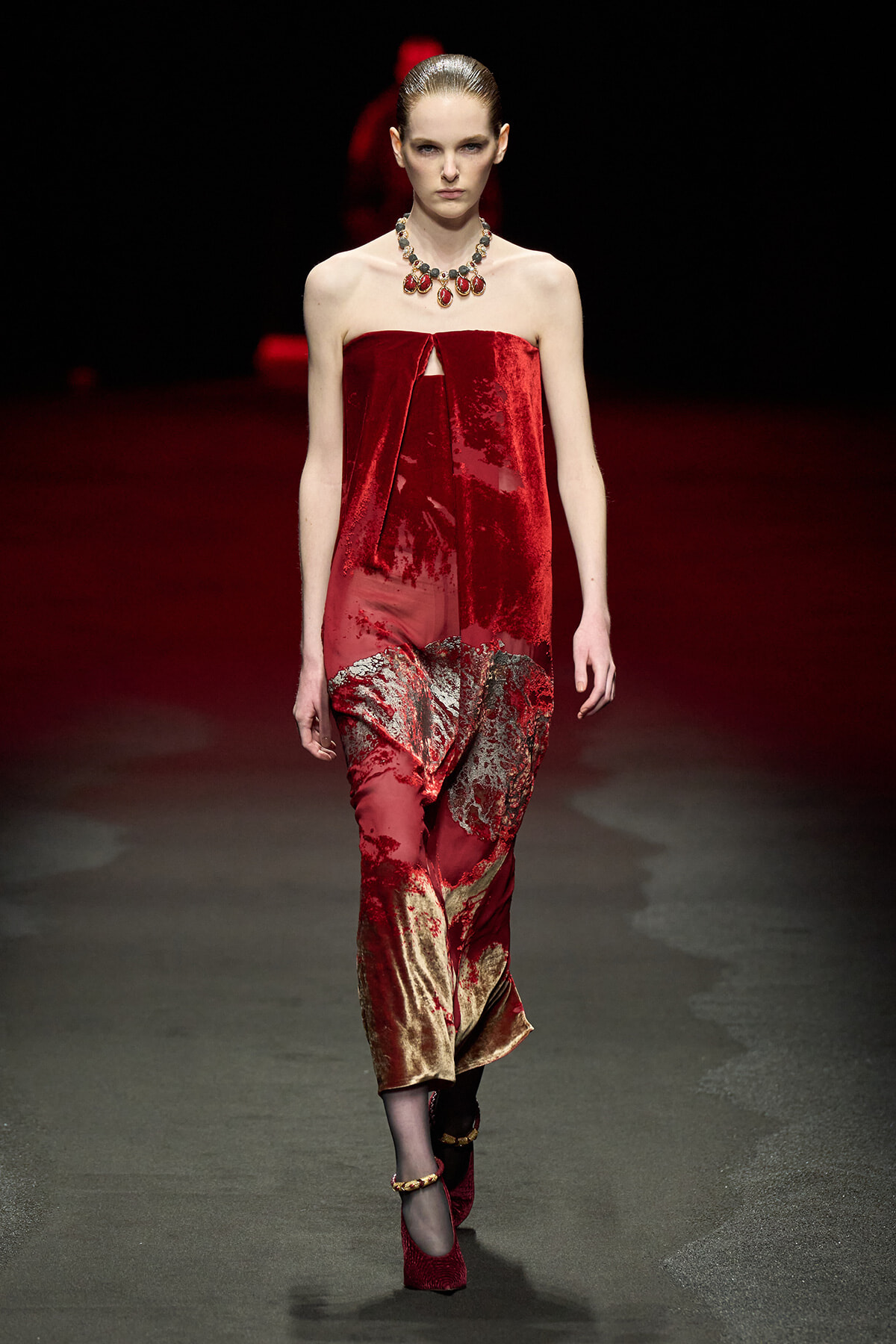 Model on a runway wearing a strapless red velvet dress with a draped, metallic gold-red skirt and a statement red gemstone necklace.