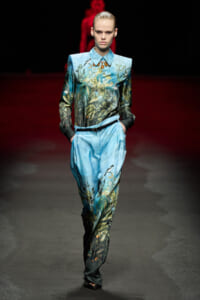 Model walks the runway in a blue botanical-print coordinated top and wide-leg pants with a belt and hands in pockets on a dark stage.