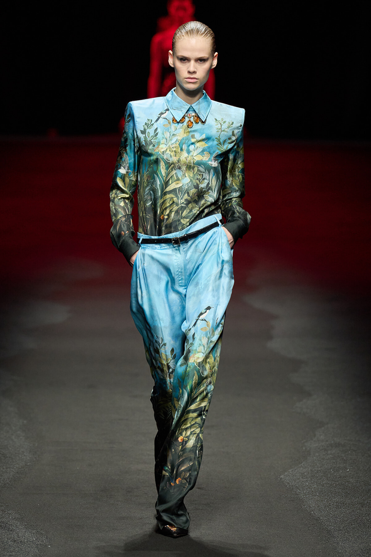 Model walks the runway in a blue botanical-print coordinated top and wide-leg pants with a belt and hands in pockets on a dark stage.