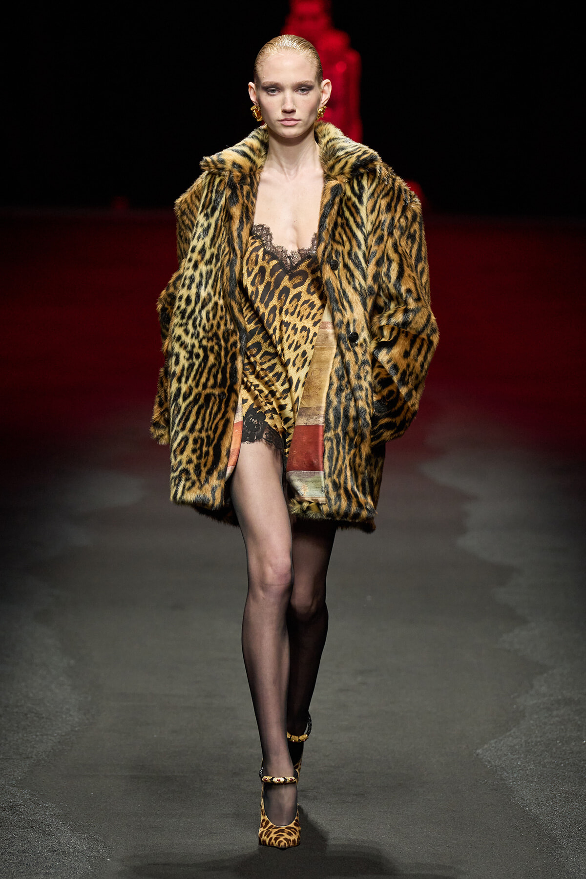 Model walks the runway in a bold leopard-print fur coat over lace lingerie and stockings, with gold earrings and leopard heels.