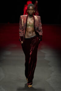 Model walks a runway in a burgundy velvet suit: textured blazer, gold bralette, and leopard-print shoes; dramatic dark backdrop enhances the ensemble.