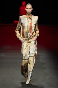 Model walking a fashion runway in a beige–earth-toned, abstract patchwork jumpsuit with a belt and leopard-print shoes, hands in pockets, spotlighted against a dark red background.