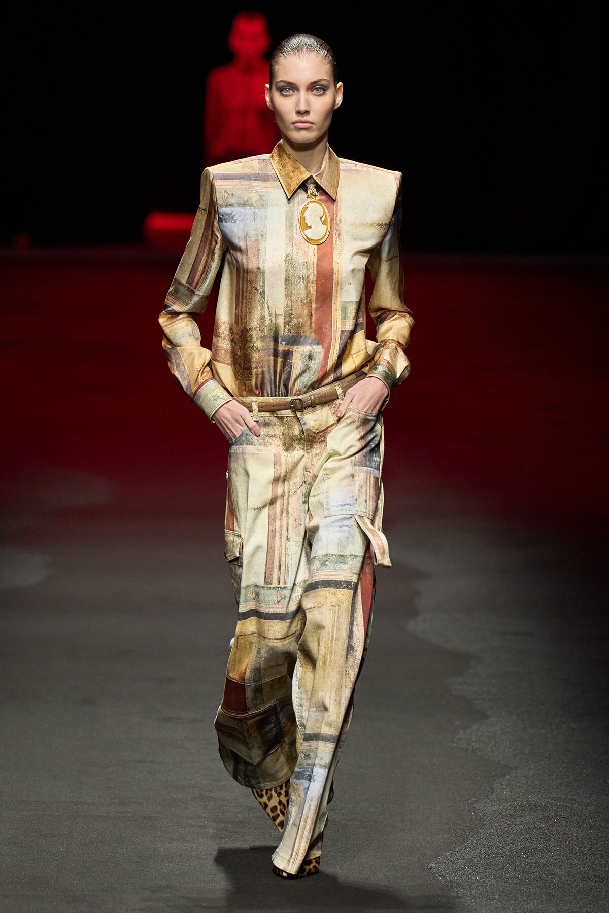 Model walking a fashion runway in a beige–earth-toned, abstract patchwork jumpsuit with a belt and leopard-print shoes, hands in pockets, spotlighted against a dark red background.