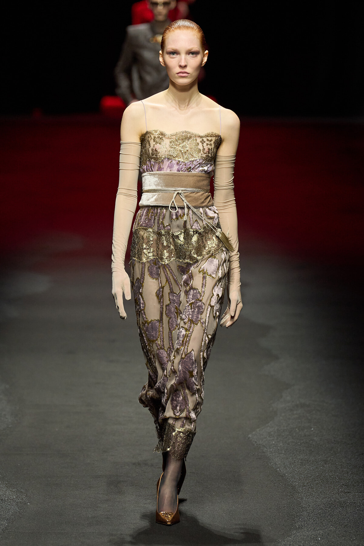 Model on a runway wearing a lavender-gold lace evening gown with sheer beige sleeves and a wide satin belt.