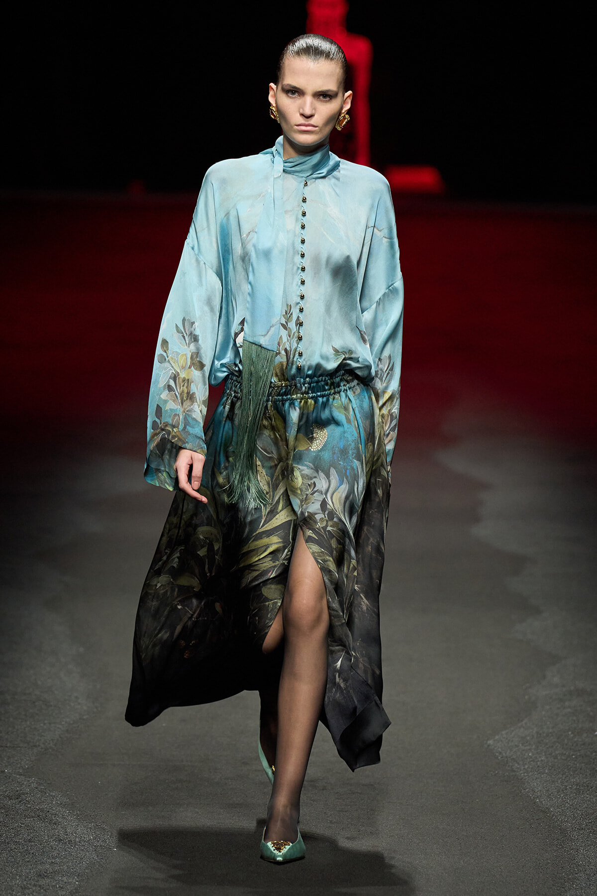 Model walks a runway in a teal-blue, long-sleeve satin dress with botanical print and a high front slit, accessorized with gold earrings and teal flats.