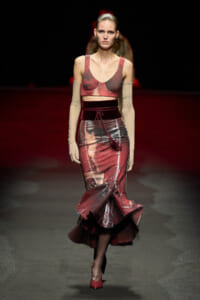 Runway model wearing a red-toned cropped top and glossy mermaid-style skirt with a velvet belt and tassel, on a dark catwalk.