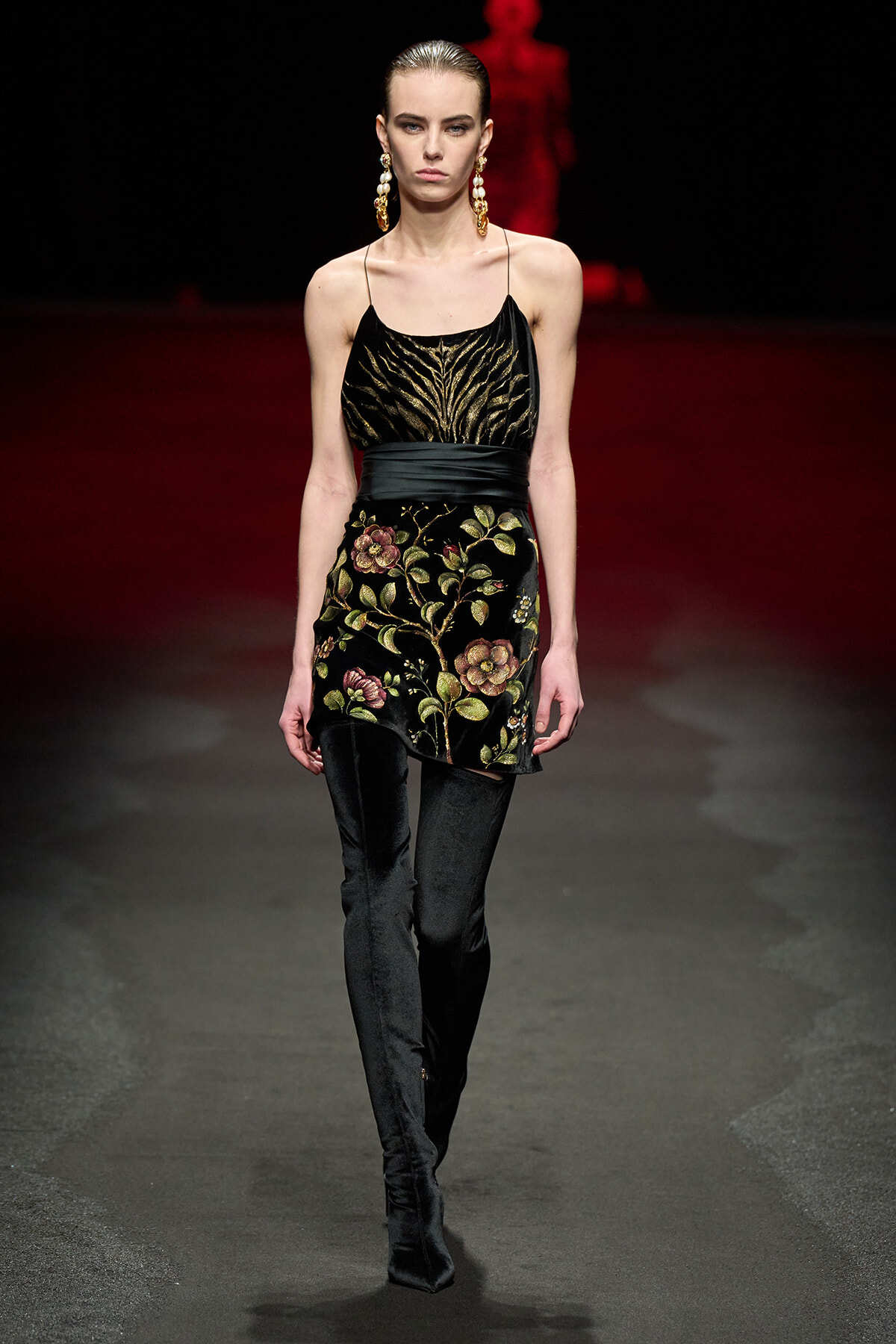 Model walks the runway in a black velvet mini dress with gold floral embroidery and matching thigh-high boots.