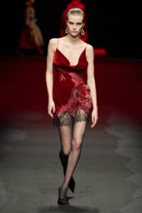 Model on a fashion runway wearing a red velvet slip dress with a lace-trimmed hem and sheer black stockings.
