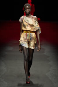 Runway model wearing a one-shoulder, abstract-print romper with a mustard belt and red heels.