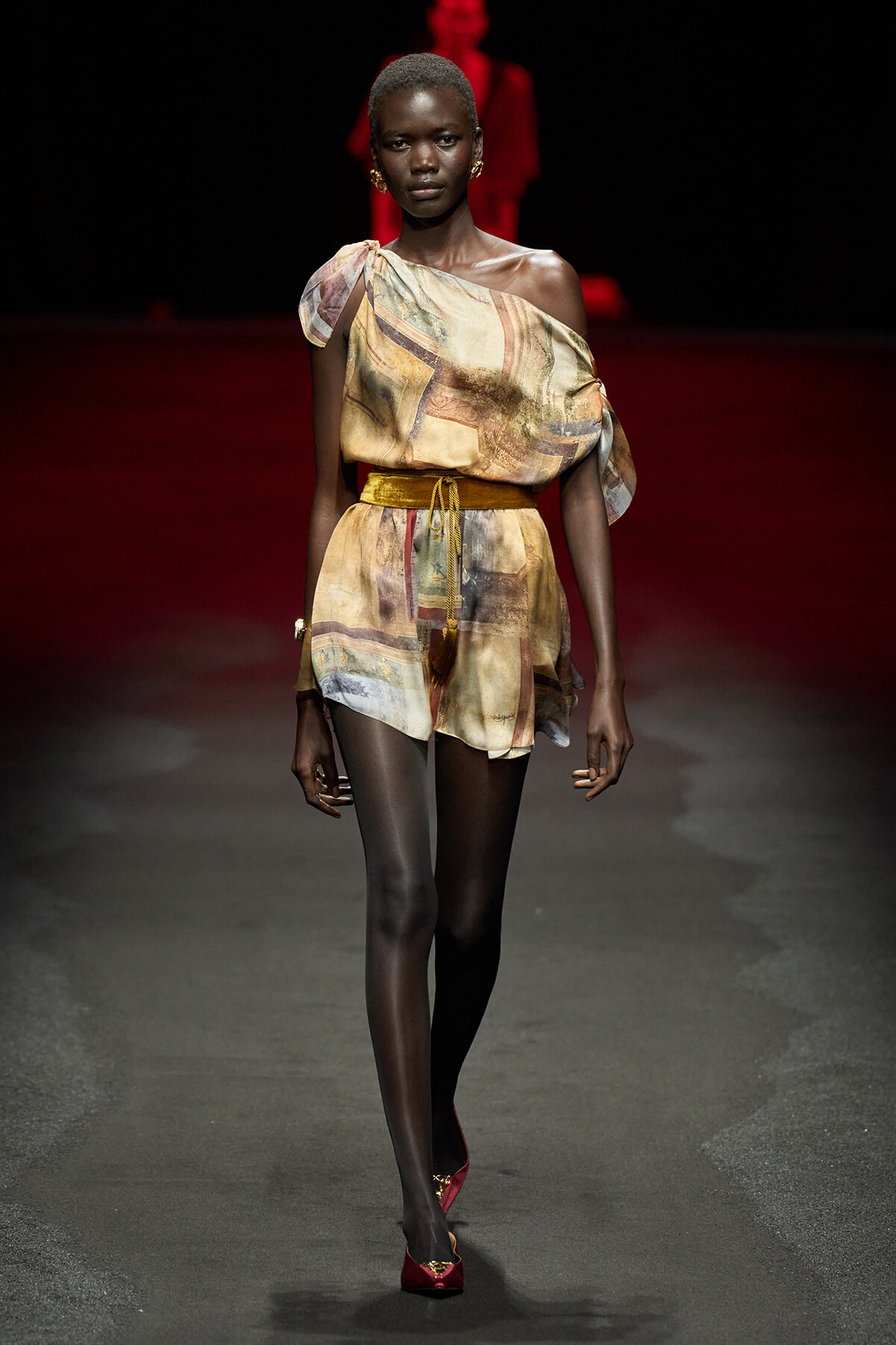 Runway model wearing a one-shoulder, abstract-print romper with a mustard belt and red heels.