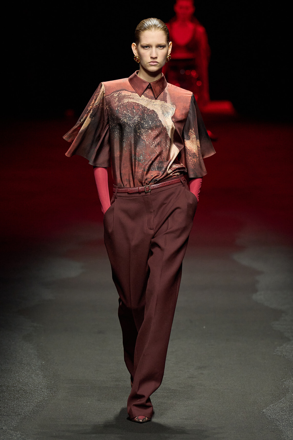 Runway model in burgundy satin blouse with abstract print and matching high-waisted trousers, red gloves, and gold earrings.