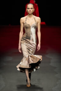 Model walking the runway in a metallic champagne mermaid gown with a structured halter neckline and a dramatic flared hem.