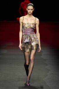 Model walks the runway in a sheer gold lace dress with floral appliqués and a wide mauve belt with tassels, paired with purple heels.