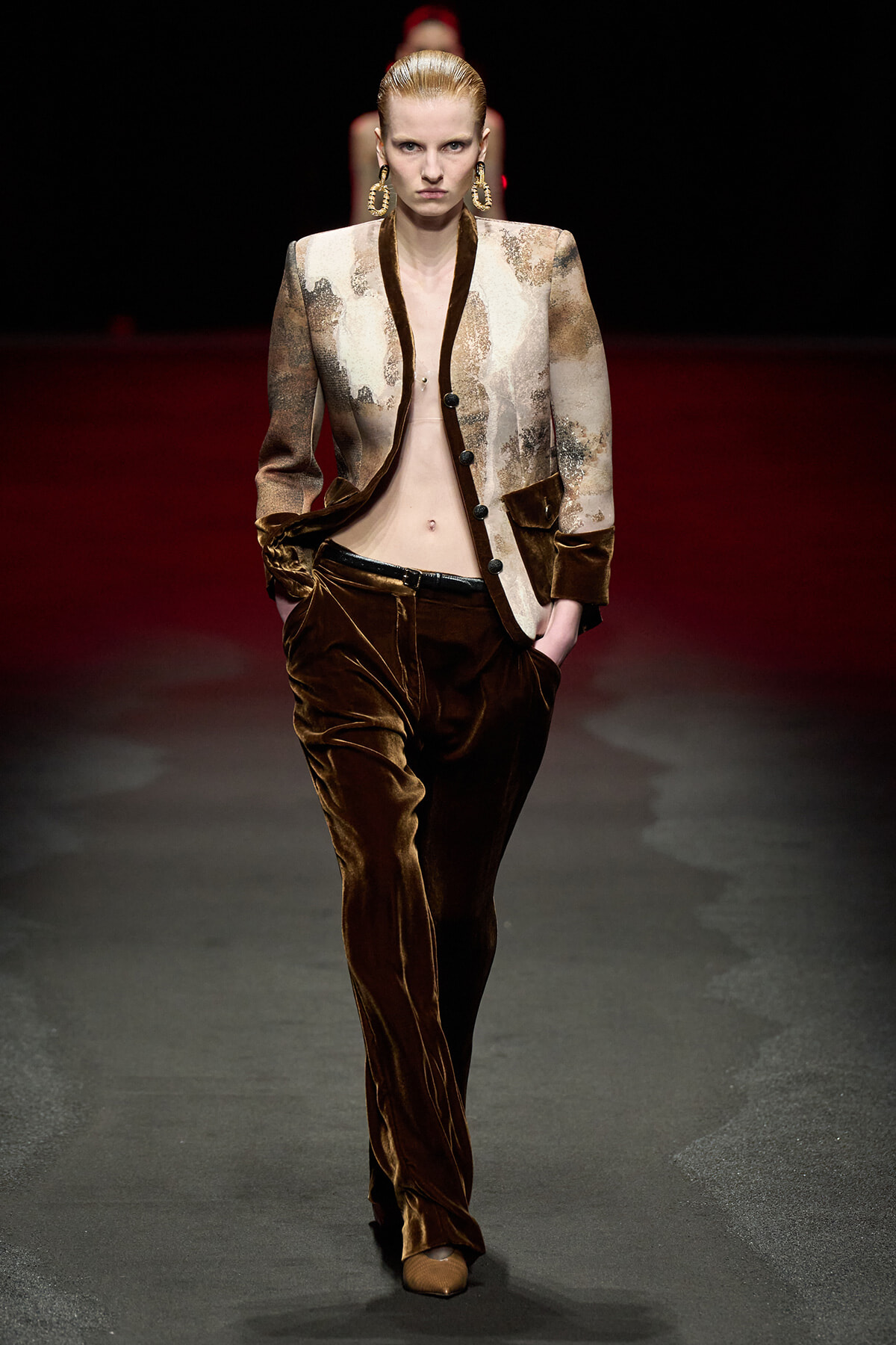 Model on a dark runway wearing a two-tone patterned blazer with brown velvet lapels and matching brown velvet trousers, hands in pockets, bold gold hoop earrings.