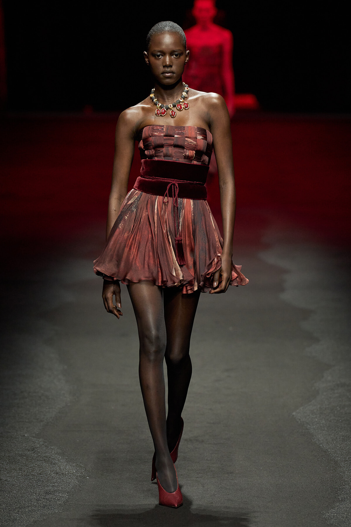 Model walking a runway in a strapless burgundy dress with a layered tulle skirt, velvet belt, red heels, and a bold statement necklace.