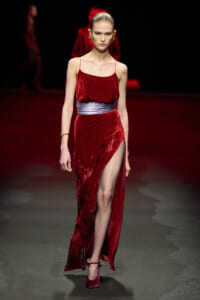 Model walking the runway in a red velvet slip dress with a silver sash and high slit.